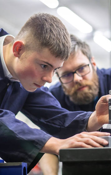A lecturer and a student in navy-blue overalls working together in a mechanical engineering workshop. The lecturer is demonstrating how to use a tool on a metal component held in a vise. A lecturer and a student in navy-blue overalls working together in a mechanical engineering workshop. The lecturer is demonstrating how to use a tool on a metal component held in a vise.